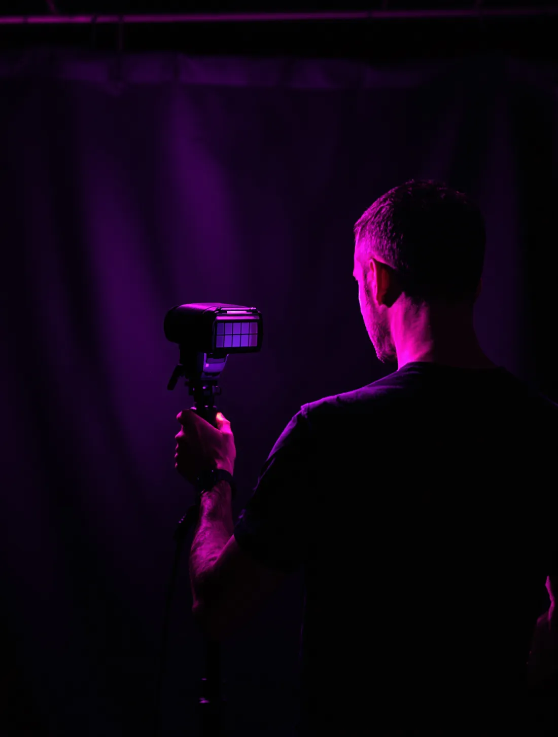Elliot from behind, adjusting a gridded speedlight with a magenta gel in a dark studio.